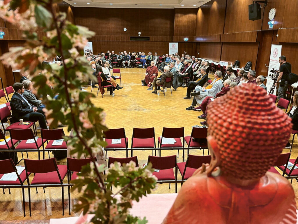 Bild einer Diskussionsrunde mit Forschenden, Ordinierten und Leitenden aus Verbänden und Gemeinschaften. Aufgenommen von oben. Ein Buddhakopf von hinten nimmt einen großen Teil des Bildes rechts unten ein. 