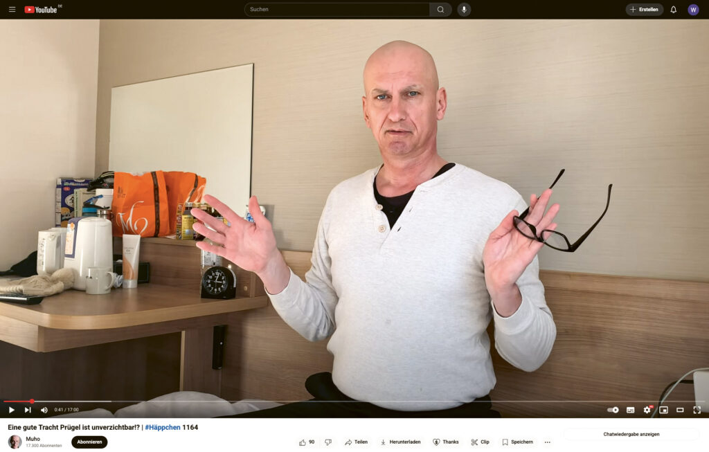 Muho in seinem YouTube-Video #Häppchen 1164 in einem weissen T-Shirt und mit einer Brille in der Hand.