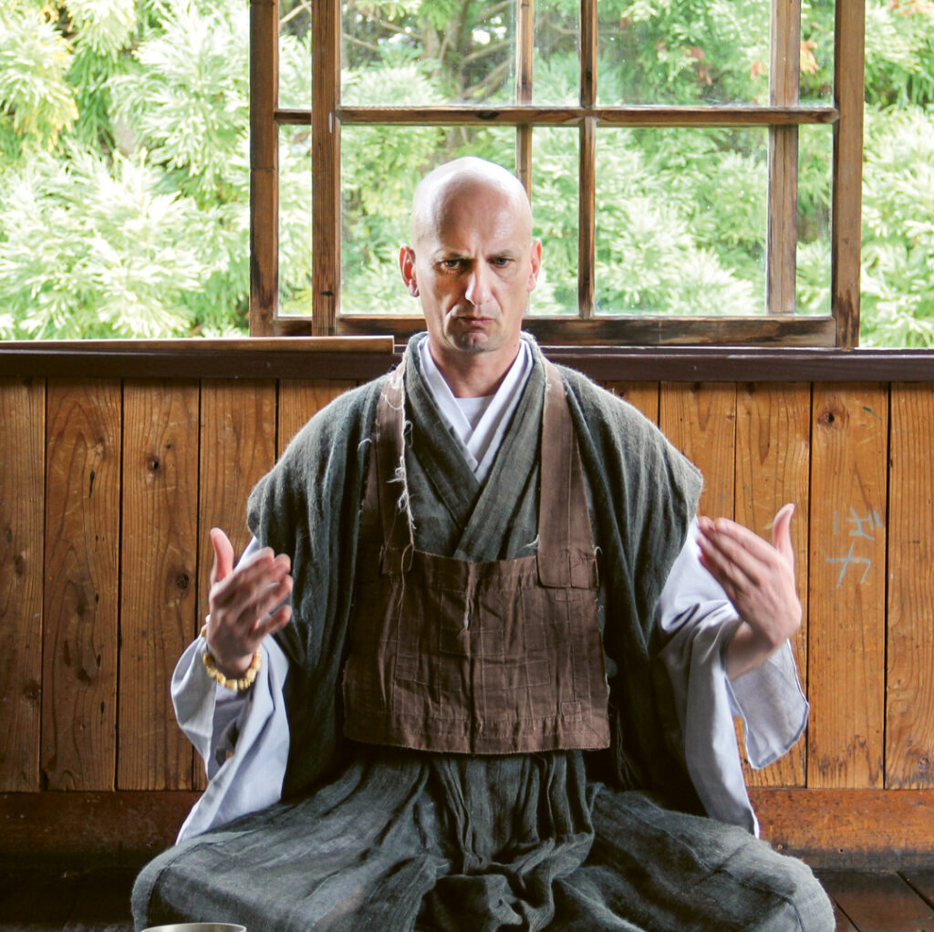 Muho unterrichtet bei einem Yoga-Event in der Präfektur Iwate. Er sitzt vor einer Holzwand im Schneidersitz.