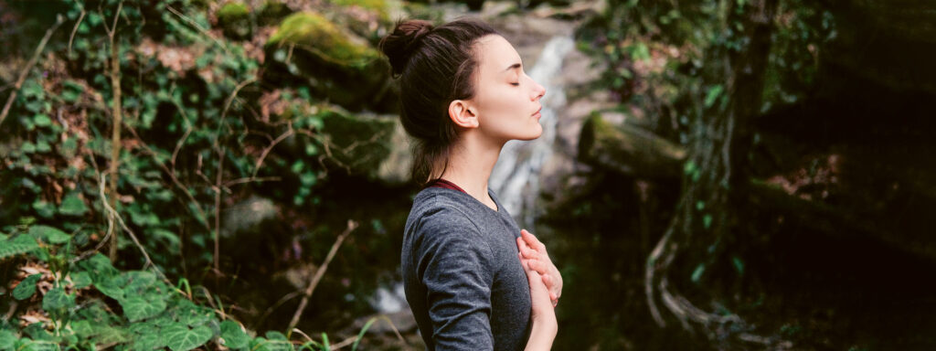 Junge Frau in der Natur, Seitenansicht, vor Wasserfall. Auge geschlossen, Hände übereinander auf der Brust. 