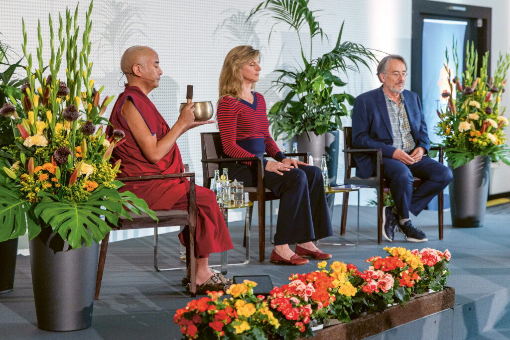 Mingyur Rinpoche, Maja Göpel und Gert Scobel sitzen auf einem Podium auf Stühlen, umgeben von Blumen. Mingyur Rinpoche hält eine Klangschale, alle haben die Augen geschlossen. Foto: © Artus Geisler