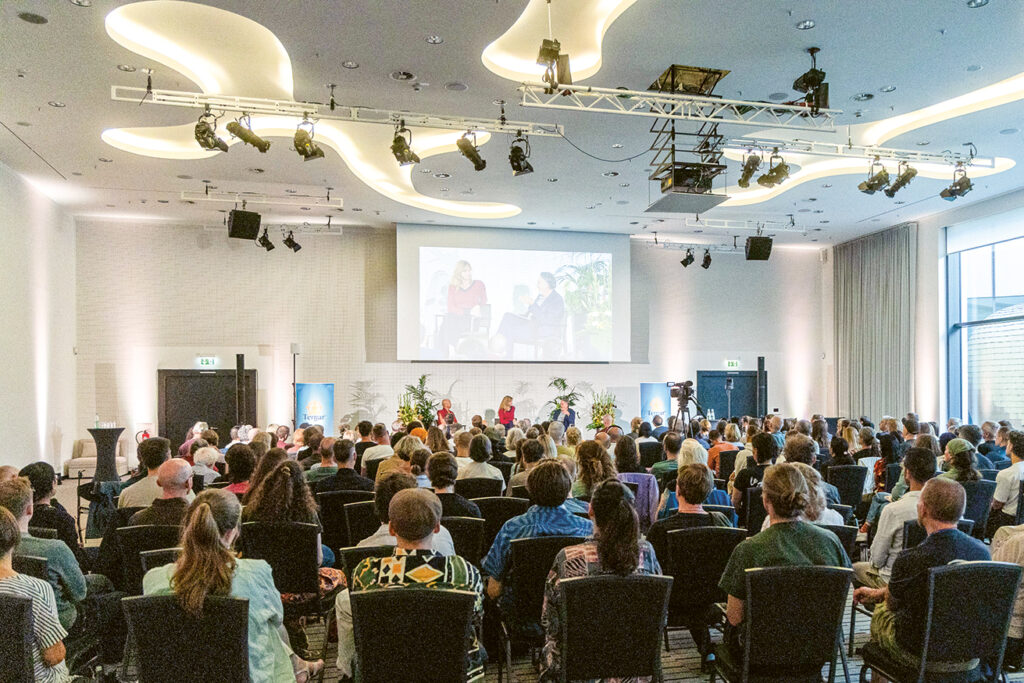 Ein großer Hörsaal it vielen Menschen auf Stühlen, Mingyur Rinpoche, Maja Göpel und Gert Scobel auf dem Podium, über eine Leinwand werden die Sprecher:innen vergrößert für alle sichtbar gemacht. Foto: © Artus Geisler 