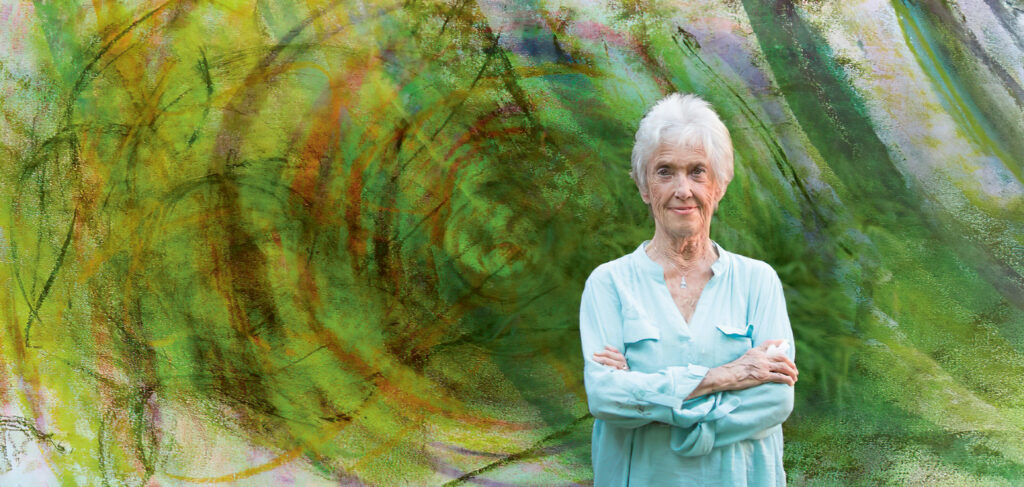 Joanna Macy vor abstrakten Gemälde in Grün-Braun-Tönen. Erkennbar sind Ringe. 